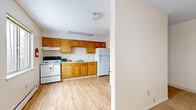 Located at 40 Court St in Geneseo, NY, this single-floor unit offers five bedrooms, one bathroom, and a well-organized layout connected by three hallways. The kitchen features warm honey-toned wood cabinetry arranged in an L-shape along one wall, complemented by white appliances including a range, refrigerator, and sink with ample counter space. Light wood-style flooring runs through the kitchen and common areas, creating a clean, cohesive look. A half-wall partition separates the kitchen from the adjacent living area, which transitions into carpet and is brightened by multiple windows fitted with horizontal blinds.

The bedrooms are carpeted and uniformly neutral in tone, each equipped with baseboard heating and windows that allow natural light. Select bedrooms include closets with bi-fold louvered doors. The shared bathroom features light wood-style flooring, a tiled tub-shower combination, a toilet, and a window for natural ventilation. Baseboard heating is present throughout the unit, and a wall-mounted air conditioning unit provides cooling in the main living area. The textured ceilings and neutral wall tones give the entire space a consistent, practical aesthetic well-suited for multiple occupants.