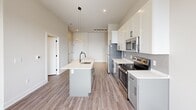 Located at 340 S Market St in Wilmington, Delaware, this two-bedroom, one-bathroom unit offers a well-designed single-floor layout with a clean, contemporary aesthetic. The open-concept kitchen serves as the centerpiece of the living area, featuring a central island with a built-in sink and matte black faucet, crisp white upper cabinetry, contrasting gray lower cabinets, white quartz countertops, and a full suite of stainless steel appliances including a refrigerator, electric range, and microwave. Pendant lighting and a track light fixture illuminate the space, while wood-look flooring ties the kitchen and living area together seamlessly. Large windows allow natural light to fill the open living space, enhancing the bright, neutral palette throughout. Both bedrooms are carpeted and finished with soft neutral tones; the primary bedroom offers a comfortable footprint, while the secondary bedroom features large windows that invite ample daylight. A dedicated closet with built-in shelving provides organized storage. The bathroom includes a glass-enclosed shower/tub combination with black-framed sliding doors and wood-look tile flooring, completing the modern design. Overall, the unit presents a cohesive, contemporary living environment suited for downtown Wilmington living.
