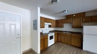 This single-floor space offers a practical and well-organized layout designed to meet everyday living needs. The kitchen is a standout feature, showcasing warm honey-toned wood cabinetry that extends to the upper and lower walls, providing generous storage throughout. The kitchen is equipped with a white electric range and oven, an under-cabinet range hood, and a double-basin sink set beneath additional upper cabinetry. Dark laminate countertops contrast nicely with the lighter wood tones, adding visual depth to the space. The flooring throughout features light wood-look vinyl planking, lending a clean and cohesive look that is both durable and easy to maintain. The ceiling is finished in white with flush-mounted lighting fixtures and a central ventilation vent, keeping the space feeling bright and airy. A structural column and an electrical panel are thoughtfully integrated near the kitchen entry, reflecting the utilitarian design of the space. The neutral white walls provide a versatile backdrop, making the space easy to personalize. Overall, the layout is efficient and functional, ideal for those seeking a straightforward, move-in-ready space.
