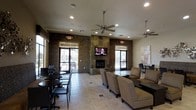 This single-floor community lounge is designed to provide a comfortable and functional gathering space for residents and guests alike. The room's most striking architectural feature is a floor-to-ceiling stacked stone fireplace, which anchors the main wall and serves as a natural focal point. A flat-screen television is mounted directly above the fireplace, making the area ideal for relaxed viewing. The walls are accented with rich, dark stone panels that lend a warm, contemporary character to the interior. Light tile flooring runs throughout the space, contributing to a clean and open aesthetic. Recessed lighting, complemented by two sleek ceiling fans with pendant-style fixtures, ensures the room is well-lit and comfortable across all seasons. The layout accommodates a variety of seating arrangements, including patterned upholstered armchairs grouped around a central coffee table for casual conversation, as well as smaller bistro-style tables with dark chairs positioned near the windows for more intimate or task-oriented use. Large glass doors on either side of the fireplace wall open directly to an outdoor patio area, flooding the interior with natural light and providing a seamless connection to the exterior. The overall ambiance is inviting and versatile, suited for both social gatherings and quiet individual use.