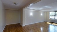 Located at 675 N Terrace Ave in Mount Vernon, NY, this single-floor space offers a clean, functional layout with a welcoming and neutral aesthetic. The interior is defined by rich hardwood flooring that extends throughout, showcasing a warm tonal variation and a decorative inlay border detail that adds a refined touch to the open living area. Walls are finished in a soft, neutral tone that reflects light and contributes to an airy atmosphere. Wall-mounted sconce lighting provides a warm, ambient glow across the main living space, complementing the overhead flush-mount ceiling fixture. A structural column element divides portions of the space while maintaining an open, connected feel between areas. White painted baseboards provide clean contrast against the hardwood floors, and the overall layout suggests a well-organized, move-in ready interior. The space benefits from a straightforward single-level design that promotes easy flow between areas. The understated finishes and classic architectural details make this a versatile space well-suited for a variety of uses.