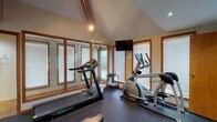 Located at 7150 Bent Tree Boulevard in Columbus, Ohio, this dedicated fitness center offers a well-equipped, single-floor workout space designed for comprehensive exercise. The cardio area features a treadmill and an elliptical machine positioned along a wall of wood-trimmed windows fitted with horizontal blinds, allowing ample natural light to fill the room while maintaining privacy. A wall-mounted flat-screen TV is conveniently positioned for viewing during workouts. The vaulted ceiling in the cardio section adds a sense of openness and volume to the space. The strength training area transitions into a lower-ceilinged zone with recessed lighting, housing a BodyCraft multi-function cable machine and an adjustable weight bench, providing a versatile setup for a full range of resistance exercises. A set of dumbbells and a water fountain are also present. Full-length mirrored walls run along one side, visually expanding the space and supporting proper form during exercise. The durable rubber flooring throughout ensures safety and comfort underfoot. The overall ambiance is functional and bright, making this fitness center a practical and well-appointed space for a complete workout routine.