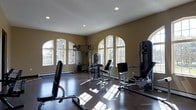 This dedicated fitness space is designed to support a comprehensive workout experience within a single-floor layout. The room is immediately distinguished by a row of elegant arched windows that line the walls, flooding the interior with generous natural light and offering views of surrounding greenery. The warm tan-toned walls and recessed ceiling lighting create a bright yet inviting atmosphere well-suited for exercise.

The polished dark hardwood flooring adds a refined aesthetic while providing a durable surface throughout the space. Multiple professional-grade strength-training machines are thoughtfully arranged across the room, including a cable pulley system, a leg press machine, an adjustable bench, and additional resistance equipment, offering a variety of upper and lower body workout options.

The room's generous proportions allow for comfortable spacing between each piece of equipment, reducing any sense of clutter and enabling free movement during exercise. Baseboard heating is visible along the perimeter, ensuring year-round comfort. Overall, this fitness room combines functional design with classic architectural details, making it a well-appointed space for dedicated personal training.