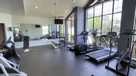 This fitness center is designed to support a well-rounded workout experience within a single-level layout. The space features vaulted ceilings that create an open, airy atmosphere, enhanced by a ceiling-mounted fan that promotes air circulation during workouts. Large, multi-pane windows line one wall, flooding the room with abundant natural light and offering views of the surrounding greenery outdoors.

The cardio area is outfitted with True-brand treadmills and an elliptical machine, positioned to take advantage of the natural light and outdoor views. A dedicated free weights section includes a flat bench and a rack of dumbbells, supported by a full-length wall mirror that expands the sense of space and allows users to monitor their form. Adjacent to the free weights is a rubber-floored stretching and mat area, providing ample room for floor exercises and flexibility work.

A wall-mounted flat-screen television is positioned near the mirror, adding an entertainment element to the cardio and stretching zones. The dark rubber flooring throughout the space provides durability and safety underfoot, while the overall design balances functionality with a clean, modern aesthetic.