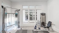 Located at 2211 Parklake Dr NE in Atlanta, Georgia, this fitness center is a well-appointed, single-level space designed to support a comprehensive workout experience. The interior features a clean, modern aesthetic with crisp white walls, recessed lighting, and a combination of light hardwood-style flooring and durable checkered rubber fitness flooring in the main exercise area.

The facility is equipped with a robust selection of Precor strength machines, a cable crossover system, and a full rack of dumbbells ranging across multiple weight increments, suitable for users of all fitness levels. A wall-mounted flat-screen television provides entertainment during workouts, while a large floor-to-ceiling mirror along one wall enhances the sense of space and allows for form monitoring. Stability and balance training accessories, including a BOSU ball and exercise ball, are also available.

Natural light filters into the space through multiple large windows fitted with roller shades, offering a bright and energizing atmosphere. Glass-paneled double doors with transom windows connect the strength training area to an adjacent cardio room, which is visible through the interior windows and contains additional treadmills and equipment. The overall design balances functionality with a polished, contemporary finish, making this fitness center a practical and well-maintained amenity.