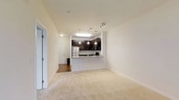 This single-floor space offers a clean, modern aesthetic defined by an open layout that promotes an airy and connected living experience. The living area is finished with plush neutral-toned carpet, contributing to a comfortable and inviting atmosphere, while crisp white walls and smooth ceilings enhance the sense of openness throughout.

The kitchen is a standout feature, showcasing rich dark espresso cabinetry that provides ample storage alongside light granite countertops. A peninsula-style breakfast bar extends into the living space, creating a natural divide while maintaining visual continuity between the two areas. Stainless steel appliances, including a refrigerator and microwave, add a polished, contemporary touch. Pendant lighting hangs above the breakfast bar, complementing the track lighting overhead that ensures the kitchen is well-illuminated.

A hallway visible from the main living area leads to additional rooms and includes hardwood-style flooring, offering a pleasing contrast to the carpeted living space. The entry door and adjacent doorways are framed with clean white trim, consistent with the overall refined and neutral design palette. The space as a whole is thoughtfully designed to balance functionality with modern style, making it both practical and visually appealing.