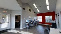 This single-floor space is defined by its striking vaulted ceilings that rise dramatically overhead, creating an airy and expansive atmosphere throughout. Natural light floods the interior through a series of skylights positioned along the roofline, supplemented by recessed fluorescent lighting that ensures consistent illumination across the open floor plan. The flooring is a warm-toned hardwood throughout, adding a refined finish to the contemporary design.

The main area serves as an open workstation hub, featuring a central desk with ergonomic mesh office chairs, ideal for individual or collaborative work. Along one wall, a row of desktop computer stations is set against a bold red accent wall framed by large windows that invite additional natural light and outdoor views. This computer station area provides ample seating and workspace for multiple users simultaneously.

Doorways on either side of the main space lead to adjacent rooms, one of which appears to be a dedicated conference or meeting room furnished with a table and chairs, offering a more private setting for group discussions. The walls are painted in cool neutral gray tones, complementing the clean, professional aesthetic. A vibrant abstract artwork adds a dynamic focal point to the main area, enhancing the overall modern and welcoming character of the space.