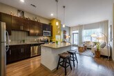 This thoughtfully designed single-floor residence offers a seamless blend of modern aesthetics and functional living. The heart of the home is the open-concept kitchen, which boasts rich dark cabinetry, a sleek tile backsplash, stainless steel appliances, and a generous granite-topped island with bar seating — ideal for casual dining or entertaining. Pendant lighting above the island adds a warm, inviting glow, while hardwood-style flooring flows throughout the main living area.

The living room is bright and airy, benefiting from large windows dressed with sheer curtains that filter in abundant natural light and offer views of surrounding greenery. A glass door provides direct outdoor access, enhancing the connection between interior and exterior.

The bedroom is a calm, comfortable retreat featuring plush carpeting, a large window with sheer drapery, and a well-organized closet. Its neutral tones and soft finishes create a serene atmosphere well-suited for rest.

The bathroom is finished with large-format white tile flooring, a dark-cabinetry vanity with a framed mirror, and a full tub-shower combination. Consistent use of a neutral gray and yellow accent palette ties the space together, giving the entire residence a cohesive, contemporary character.