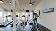 This fitness center is thoughtfully designed to accommodate a full range of workout activities within a single-floor layout. The space is divided into distinct functional zones, each catering to different exercise needs. The cardio area is lined with multiple treadmills and elliptical trainers, positioned to face large glass doors that flood the room with natural light and offer views to the outdoors. A ceiling fan overhead provides added ventilation and comfort during workouts. Full-length mirrors along the walls visually expand the space and allow users to monitor their form. A mounted flat-screen TV adds an entertainment element to the cardio zone. The strength training area features a multi-station cable machine and additional resistance equipment, set against sage green accent walls beneath a wood-paneled ceiling with track lighting. Rubber flooring throughout provides safety and durability. A large sliding glass door in the strength zone opens to an outdoor area, enhancing airflow and connectivity to the exterior. The space also includes a sanitization station and posted instructional signage, reflecting a clean and user-friendly environment. The combination of vaulted ceilings, bright white walls, and ample natural light gives the fitness center an open, energizing atmosphere.