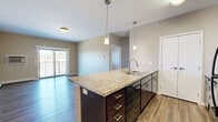 This well-appointed 2-bedroom, 2-bathroom unit at 1502 Belsly Blvd in Moorhead, MN offers a thoughtfully designed single-floor layout. The open-concept kitchen is a standout feature, anchored by a large center island with a granite-style countertop, undermount sink, and elegant pendant lighting. Dark espresso cabinetry pairs with black appliances, including a refrigerator, range, and microwave, while hardwood-style flooring ties the kitchen seamlessly into the living area. The living room is bright and airy, with a sliding glass door that opens to a private balcony, inviting natural light and outdoor views. Both bedrooms are carpeted for comfort, each featuring windows with blinds and baseboard heating. The primary bathroom offers a walk-in shower and a granite-top vanity with dark cabinetry, while the second bathroom includes a full tub-shower combo with the same refined finishes. Tile flooring runs throughout both bathrooms. A dedicated in-unit laundry room houses a full-size washer and dryer with overhead shelving for added storage. Neutral warm-toned walls and consistent finishes throughout create a cohesive and inviting atmosphere.