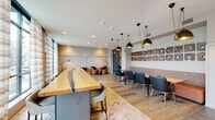 This single-floor co-working and amenity space is designed to accommodate a variety of work and social needs, blending modern aesthetics with functional versatility. The main open area features light wood-look flooring throughout, anchored by a striking live-edge communal table with a blue resin inlay, serving as a central workspace. Pendant lighting with brass and matte black finishes hang overhead, adding an upscale industrial character to the room. A built-in banquette with a warm terracotta upholstered seat runs along one wall, paired with navy dining-style chairs and dark wood tables beneath a curated arrangement of decorative wall art. A dedicated lounge nook offers comfortable cognac leather swivel chairs clustered around low tables, with a wall-mounted display screen ideal for presentations or communal use. Large floor-to-ceiling windows dressed in sheer curtains flood the space with natural light and provide views of the surrounding streetscape. A glass-walled meeting room provides a private retreat, outfitted with an elegant round conference table, leather task chairs, and a sculptural chandelier. A smaller private room features bold teal grasscloth walls, botanical art prints, and a gold chandelier, creating an intimate setting. A beverage station and kitchenette area complete the space, alongside a well-appointed entry corridor with curated artwork.