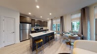 Located at 59 2nd Ave in Raritan, New Jersey, this single-floor residence offers a thoughtfully designed open-concept layout with contemporary finishes throughout. The kitchen is a standout feature, equipped with dark espresso cabinetry, stainless steel appliances including a refrigerator, microwave, and gas range, and a large center island topped with a crisp white quartz surface. Gold-accented bar stools add a stylish touch, while the adjacent dining area benefits from generous natural light streaming through floor-to-ceiling windows dressed with layered roller shades and grommet curtains. Light wood-style flooring flows seamlessly across the main living areas, enhancing the sense of openness. The space includes two well-proportioned bedrooms, each featuring large windows with outdoor views of lush greenery, providing a tranquil atmosphere. One bedroom offers direct access through a set of full-height windows, while the other benefits from corner windows that maximize natural light. The bathroom is finished with neutral tile, a soaking tub and shower combination, a dark wood vanity with a white countertop, and a frameless mirror. Recessed lighting throughout the home ensures a clean, modern aesthetic. Overall, the residence combines functional design with refined details in a well-lit, inviting environment.