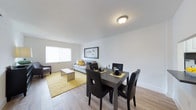 This single-floor space is designed with an open-concept layout that seamlessly connects the living and dining areas, creating a comfortable and functional environment. The living room features light wood-style flooring that runs throughout, white walls, and a large window that allows natural light to fill the space. A grey sofa anchors the seating area, complemented by a sleek metallic coffee table and a vibrant yellow geometric area rug that adds a pop of color. A framed abstract artwork on the wall and a dark wood credenza with a decorative lamp complete the living area's contemporary aesthetic. Adjacent to the living room, the dining area accommodates a dark wood dining table with four black upholstered chairs, making it well-suited for everyday meals or entertaining guests. The open sightlines between the two areas enhance the sense of spaciousness throughout. Clean white walls and simple flush-mount ceiling lighting maintain a bright, airy atmosphere. The consistent flooring and cohesive color palette of grey, black, and yellow tie the entire space together, resulting in a modern, inviting interior that balances style with practicality.