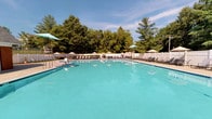 This outdoor aquatic facility offers a thoughtfully designed recreational environment centered around a generously sized swimming pool with clear turquoise water. The pool deck is lined with lounge chairs and teal patio umbrellas, providing ample space for relaxation in the sun. A white perimeter fence encloses the area, enhancing privacy while mature trees form a natural green backdrop.

Adjacent to the pool, a covered patio area extends the usable space and features natural slate flagstone flooring in earthy blue-grey tones, adding a refined touch to the outdoor setting. The patio is furnished with sling-style lounge chairs and round metal tables, making it an ideal spot for poolside gatherings or shade-seeking guests. Structural columns support the covered roof, creating a comfortable semi-sheltered retreat.

Elegant French doors with white-painted frames connect the covered patio to an interior space, with large glass panes offering views of the pool from within. A brick building with a service window overlooks the pool deck, suggesting the presence of support amenities. Access to the pool area is managed through a stately black wrought iron security gate with decorative finials, reinforcing the sense of a well-maintained, secure facility. Overall, the space strikes a balance between functional amenity and polished outdoor design.