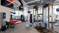 This modern fitness center is designed to support a wide range of workout styles, combining cardio, strength, and functional training in one cohesive space. Large floor-to-ceiling windows line the perimeter, flooding the interior with natural light and offering views of tropical palm trees and lush landscaping, creating an energizing atmosphere for exercise. The elevated ceilings, finished in a deep charcoal tone, add a sense of spaciousness while lending the space a polished, contemporary aesthetic. Linear lighting fixtures provide consistent illumination throughout. The cardio zone features a row of treadmills positioned along the windowed wall, ideal for scenic workouts. The strength and functional training area is well-appointed with a multi-functional cable and power rack, a suspended heavy punching bag, kettlebells, battle ropes, and weighted resistance rollers. Wall-mounted flat-screen televisions are strategically placed throughout for entertainment. Full-length mirrors enhance the sense of depth, and durable, patterned carpet tiles cover the floor, providing both comfort and practicality. The overall ambiance is sleek, modern, and motivating.