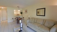 This single-floor space offers a thoughtfully designed living area that balances comfort and style. The living room immediately draws attention with its soaring ceilings, which lend an open and airy feel to the room. Natural light streams in through a large window dressed with a Roman shade, as well as a glass-paneled door that provides access to the exterior while brightening the interior throughout the day. The walls are finished in a soft, neutral tone that creates a calm and welcoming ambiance. A patterned sofa with decorative throw pillows serves as the focal point of the seating area, complemented by a set of ornate, dark metal drum-style accent tables that add a touch of eclectic character. A traditional area rug anchors the space, while two classically framed art pieces displayed side by side on the wall contribute a refined aesthetic. A potted plant near the entrance adds a natural, refreshing element. The overall atmosphere is warm, intimate, and carefully curated, making this space both functional and visually appealing for everyday living.