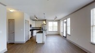This single-floor space showcases a thoughtfully designed open-concept layout that seamlessly connects the kitchen and living area. The kitchen is outfitted with crisp white cabinetry, dark countertops, and stainless steel appliances, including a refrigerator, oven, and microwave. A central island with a built-in sink serves as both a functional workspace and a natural dividing point between the kitchen and the adjoining living area. Pendant lighting hangs above the island, complementing the track lighting installed throughout the space. The living area is generously proportioned, offering ample room for furnishings and everyday living. Large windows along the far wall and a glass-panel door flood the interior with natural light, creating a bright and airy atmosphere. The consistent hardwood-style flooring runs throughout, lending a cohesive and polished look to the entire space. Neutral-toned walls enhance the sense of openness, while the clean architectural lines and modern finishes contribute to a contemporary aesthetic that is both practical and visually appealing.