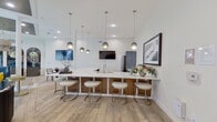 This sophisticated single-floor amenity lounge is designed with a refined, contemporary aesthetic that blends comfort with elegance. Light wood-look flooring laid in a herringbone pattern anchors the open-plan layout, which flows seamlessly across several distinct social zones.

At the heart of the space is a striking curved bar counter topped with a white marble surface and warm wood cabinetry, accompanied by sleek upholstered stools and a cluster of pendant lights overhead. Wall-mounted televisions provide entertainment throughout. A dedicated beverage and refreshment station with a glass-front refrigerator and coffee service is tucked near French glass doors leading to an adjoining room.

The lounge areas showcase rich blue velvet seating, floral-print sofas, and a plush sectional set against a bold textured accent wall finished in a sculptural tile pattern. A shimmering tiered chandelier draws the eye upward in the sitting area, where floor-to-ceiling windows dressed in sheer and patterned drapes frame lush outdoor greenery. Arched wall niches with decorative art panels add architectural character to the dining area, which features a round table and bentwood chairs.

A billiards table rounds out the entertainment offerings, completing a space designed for gathering, relaxing, and socializing in style.