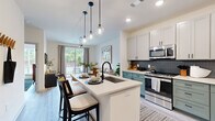This thoughtfully designed two-bedroom, one-bathroom residence offers a cohesive modern aesthetic across its single floor. Light wood-toned flooring runs throughout, complementing the crisp white walls and creating a clean, contemporary feel.

The kitchen serves as a focal point of the space, showcasing a two-tone design with white upper cabinetry and bold teal lower cabinets, accented by a white subway tile backsplash. A central island with a white countertop and undermount sink provides additional workspace and storage, while stainless steel appliances and globe pendant lighting add polished, functional detail.

The open layout connects the kitchen seamlessly to the living area, which benefits from large windows that invite abundant natural light and offer views of surrounding greenery. A distinctive sliding barn door with black hardware adds an architectural accent to the hallway.

The primary bedroom features a ceiling fan and a wall of tall windows framing outdoor views, creating an airy, light-filled retreat. The second bedroom is compact and well-proportioned, with direct hallway access.

The bathroom is well-appointed with a wood-toned vanity, white quartz countertop, framed mirror, matte black fixtures, and a glass-enclosed walk-in shower with white tile surround. A dedicated closet completes the unit's layout.