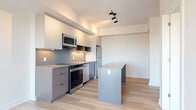 Located at 280 Montgomery St in Ottawa's Vanier neighbourhood, this well-appointed one-bedroom, one-bathroom unit offers a thoughtfully designed layout across a single floor. The open-concept living, dining, and kitchen area forms the heart of the space, featuring light hardwood flooring that extends throughout. The modern kitchen is outfitted with stainless steel appliances including a refrigerator, stove, and over-the-range microwave, complemented by two-tone cabinetry — crisp white upper cabinets paired with grey lower cabinets — a subtle tile backsplash, and a central island with a stone-look countertop that provides additional prep and dining space. Track lighting overhead adds a contemporary touch. The bedroom is a bright, quiet retreat with a large window offering outdoor views, and features a full-width mirrored sliding closet. The bathroom is finished with a clean, modern aesthetic, showcasing floor-to-ceiling white subway tile in the walk-in shower, matte black fixtures, a stone countertop vanity with dark wood cabinetry, and a glass shower panel. Additional conveniences include in-unit laundry, a hallway with two closets, and crisp white walls throughout that contribute to a bright, airy ambiance.
