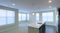 This single-floor space showcases an open-concept layout that seamlessly connects the kitchen and living areas. The kitchen features a large central island with a white quartz-style countertop and an undermount sink, complemented by dark cabinetry and modern appliances. Two pendant lights hang above the island, providing focused task lighting while adding a contemporary design touch. Recessed ceiling lights supplement the overall illumination throughout the space.

The living area extends naturally from the kitchen, featuring neutral-toned carpeting that contrasts with the hardwood flooring in the kitchen zone. Walls are finished in a soft, warm gray tone, contributing to a clean and welcoming ambiance. Multiple windows fitted with white horizontal blinds line the walls, allowing abundant natural light to fill the space from several angles. White baseboards and trim provide crisp definition throughout. The open flow between the kitchen and living area makes the space feel expansive and versatile, well-suited for both everyday living and entertaining.