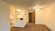 This single-floor residential space offers a practical and welcoming layout designed for comfortable living. The open-concept design seamlessly connects the kitchen and living area, creating a sense of openness and ease of movement throughout the space. The kitchen features white cabinetry, a built-in range with a black cooktop, a white refrigerator, and a peninsula with a built-in sink that acts as a natural divider between the cooking and living zones. The white cabinetry and light-colored countertops contribute to a bright, clean aesthetic. The living area is finished with wall-to-wall carpeting in a neutral tone, adding warmth underfoot and a soft, comfortable feel to the room. A ceiling fan with a built-in light fixture is centrally positioned in the living area, providing both air circulation and ambient lighting. The walls are finished in a soft, warm white tone that complements the overall palette of the space. A solid wood interior door is visible from the living area, leading to an adjacent room, suggesting a thoughtful and functional floor plan. The overall ambiance is cozy and understated, making it a practical and inviting everyday living environment.