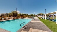 Located at 1918 W 4100 S in West Valley City, Utah, this outdoor amenity space is a well-appointed recreation area designed for relaxation and social enjoyment. The centerpiece is a generously sized swimming pool with a sleek, modern surround finished in large-format pavers, providing ample space for lounging along the pool deck. Adjacent to the pool sits a dedicated spa with clearly marked depth indicators, offering a more intimate water experience with its own entry rails and seating ledges.

The surrounding deck is thoughtfully furnished with rows of sling-style lounge chairs, perfect for sunbathing beneath Utah's expansive blue skies. A series of white pergola structures lines one side of the space, each outfitted with additional seating and providing shaded retreats from the sun. Manicured green lawn panels are interspersed throughout, adding a natural, park-like quality to the environment.

A single-story amenity building anchors one end of the space and features an outdoor dining area shaded by market umbrellas with patio table seating. Ornamental grasses and young trees are planted throughout the perimeter, offering both aesthetic appeal and a natural buffer. Views of the surrounding Utah mountain range are visible in the background, enhancing the open, airy atmosphere of this vibrant outdoor community space.
