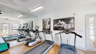 This single-level fitness center is designed to support a comprehensive workout experience, offering a thoughtfully arranged layout with a diverse selection of equipment. The space features warm hardwood-style flooring throughout, crisp white walls, and a textured ceiling with fluorescent lighting that ensures the room is well-lit at all times.

The cardio zone is anchored by multiple TRUE-brand treadmills and elliptical machines, positioned in front of a large floor-to-ceiling wall mirror that visually expands the space and allows users to monitor their form. A wall-mounted flat-screen television is positioned nearby for entertainment during workouts.

The strength training area includes a three-tier dumbbell rack stocked with a full range of hex weights, a multi-function cable machine, and a workout bench, providing options for both free weight and machine-based resistance training. A dedicated rack holds an assortment of medicine balls and stability/exercise balls in varying sizes, catering to functional and core training needs.

Natural light filters in through large windows and French doors, which offer views of an outdoor pool area. The posted fitness center rules ensure a structured, shared-use environment. Bold black-and-white athletic artwork accents the walls, contributing to an energizing and motivating atmosphere throughout the space.