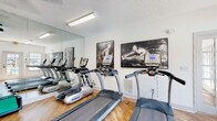 This single-level fitness center is designed to support a comprehensive workout experience, offering a thoughtfully arranged layout with a diverse selection of equipment. The space features warm hardwood-style flooring throughout, crisp white walls, and a textured ceiling with fluorescent lighting that ensures the room is well-lit at all times.

The cardio zone is anchored by multiple TRUE-brand treadmills and elliptical machines, positioned in front of a large floor-to-ceiling wall mirror that visually expands the space and allows users to monitor their form. A wall-mounted flat-screen television is positioned nearby for entertainment during workouts.

The strength training area includes a three-tier dumbbell rack stocked with a full range of hex weights, a multi-function cable machine, and a workout bench, providing options for both free weight and machine-based resistance training. A dedicated rack holds an assortment of medicine balls and stability/exercise balls in varying sizes, catering to functional and core training needs.

Natural light filters in through large windows and French doors, which offer views of an outdoor pool area. The posted fitness center rules ensure a structured, shared-use environment. Bold black-and-white athletic artwork accents the walls, contributing to an energizing and motivating atmosphere throughout the space.
