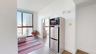 Located at 229 Duffield Street in the heart of Downtown Brooklyn, this 2-bedroom, 1-bathroom residence makes efficient use of its compact footprint across a single floor. The living room is a standout space, anchored by expansive floor-to-ceiling windows that flood the interior with natural light and frame impressive elevated views of the Brooklyn and Manhattan skyline. Light wood-style flooring runs throughout, lending warmth and continuity to the open layout. The kitchen is efficiently integrated into the main living area, featuring stainless steel appliances including a refrigerator and microwave. The primary bedroom offers a calm, airy atmosphere with a window bringing in additional natural light and views of the surrounding cityscape, complemented by a built-in desk and storage unit. The second bedroom is equally well-appointed, with clean white walls and its own window. The shared bathroom is generously proportioned, featuring large-format tile flooring, a floating vanity with a wide mirror, and a walk-in shower area. A hallway and closet complete the layout, providing practical storage solutions. Throughout the residence, crisp white walls and clean architectural lines create a modern, cohesive aesthetic well-suited to urban living in one of Brooklyn's most connected neighborhoods.