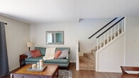 This well-organized three-floor home offers a practical and comfortable layout across its three levels. The main floor features an open-plan living and dining area bathed in natural light from a large window with dark curtains. The living space includes warm hardwood-style flooring, a carpeted staircase with white balusters and a dark handrail leading to the upper level. The kitchen is positioned just off the hallway and dining area, featuring classic oak cabinetry, dark countertops, black appliances including a refrigerator and range, and a double stainless steel sink beneath a window. The dining area connects seamlessly to the kitchen through an arched opening. Upstairs, three bedrooms are accessible via a hallway, including a generously sized primary bedroom. The upper level also features carpeted flooring, lending a quieter, more comfortable feel. Two bathrooms serve the home, finished in neutral tones with clean fixtures. The basement level offers a wide-open unfinished space with a painted concrete floor, exposed ceiling infrastructure, and a small window, presenting excellent potential for storage or future finishing. Overall, the home maintains a clean, neutral aesthetic throughout its three floors.