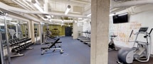 This fitness center offers a comprehensive workout environment designed to accommodate a wide range of exercise routines. The space features an open, industrial aesthetic with exposed ceiling infrastructure, including visible ductwork and piping painted white, which contributes to the room's airy, loft-like character. Concrete structural columns punctuate the floor plan, adding to the raw architectural appeal.

The cardio zone is lined with a row of treadmills, providing ample options for endurance training. Nearby, a stationary bike and exercise balls offer additional low-impact workout choices. On the opposite side of the space, a dedicated strength training area features a rack of free weights, adjustable benches, and cable machines positioned against a bold olive-green accent wall with a mirror, creating an energizing atmosphere for resistance workouts.

A large motivational typographic mural dominates one wall, featuring fitness-related keywords in a vibrant, multi-colored design that adds personality and energy to the space. Overhead, a mounted flat-screen television provides entertainment during workouts. The flooring throughout is a durable blue-gray rubberized surface, practical for heavy equipment and high-traffic use. The space is well-lit with a combination of fluorescent and pendant lighting, ensuring excellent visibility across all workout zones.