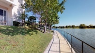 This single-floor property offers a standout outdoor feature: a beautifully maintained waterfront walkway that runs along the edge of a calm, expansive lake. The paved path is bordered by a sleek black metal safety railing on the water side, providing both security and unobstructed views across the lake to the tree-lined horizon beyond. On the landward side, mature shade trees rise from a well-kept grassy slope, creating a natural canopy that adds both character and comfort to the outdoor environment. Lush tropical and subtropical vegetation, including palms and broadleaf shrubs, frame the landscape, lending the setting a distinctly warm-climate character consistent with Florida or a similar Southern region. The staircase visible in the background suggests multi-level outdoor access, connecting upper areas of the property to the waterfront path below. The overall atmosphere is tranquil and scenic, with abundant natural light and open sky contributing to an airy, inviting ambiance. This outdoor space is ideally suited for leisurely strolls, relaxation, and enjoying peaceful water views throughout the day.