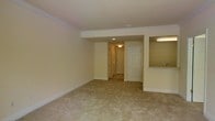 This single-floor residential space offers a practical and well-appointed layout designed for comfortable everyday living. The main living area is finished with wall-to-wall carpeting in a neutral beige tone, contributing to a warm and inviting atmosphere throughout. Crown molding lines the ceiling, adding a clean, classic architectural detail that elevates the overall finish of the space. Recessed lighting is installed in the ceiling, providing even, functional illumination across the open floor plan. The living area flows naturally toward a hallway, which leads to an exterior door — likely the main entry — finished with a traditional six-panel white door design. A mirrored closet is visible along the hallway, offering practical storage. To one side, a pass-through opening connects the living area to the kitchen, which features under-cabinet lighting and a countertop visible from the main room, creating a sense of openness between the two spaces. The walls throughout are painted in a soft, neutral cream tone, complementing the light-colored carpet and bright ceiling finishes. The overall ambiance is clean, understated, and move-in ready, with consistent finishes across the space that lend it a cohesive and well-maintained character.