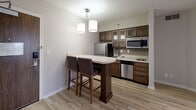Located at 115 Hutchinson Ave in Columbus, Ohio, this one-bedroom, two-bathroom unit offers a thoughtfully designed layout that maximizes every square foot of its compact footprint. The combined kitchen and living area serves as the heart of the space, featuring light-toned hardwood-style flooring, neutral walls, and a textured ceiling. The kitchen is well-appointed with rich walnut-toned cabinetry featuring glass-front upper doors, a full suite of stainless steel appliances including a refrigerator, dishwasher, microwave, and cooktop, a tile backsplash, and a sleek countertop. A peninsula breakfast bar with pendant lighting and two bar-height chairs provides a casual dining area. Sheer drape-covered windows allow natural light to filter into the living space. The bedroom offers generous proportions with patterned carpet, a bold accent wall, and a sliding wood-panel closet door alongside a secondary closet for ample storage. Both bathrooms are finished with warm-toned tile flooring, wood vanity cabinetry, granite-style countertops, and framed mirrors. One bathroom includes an accessible roll-in shower with grab bars. A utility room completes the layout, adding practical functionality to this well-organized Columbus residence.