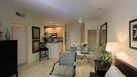 This single-floor residence showcases an open-concept design that seamlessly integrates the kitchen, dining, and living areas into one cohesive space. The kitchen features warm honey-toned cabinetry, dark countertops, stainless steel appliances including an over-the-range microwave, and a breakfast bar with a sleek barstool, providing a casual dining option alongside the more formal arrangements nearby. Recessed and track lighting throughout ensures consistent, practical illumination across the space.

The dining area is anchored by a round glass-top table surrounded by upholstered teal-toned chairs, centered with a floral arrangement that adds a refined touch. Adjacent to this, the living area is furnished with patterned armchairs in soft blue-green tones, complementing the neutral carpeted flooring that runs throughout the residence.

Framed botanical artwork adorns the walls, adding visual interest without overwhelming the light, neutral color palette. A large mirror near the living area enhances the sense of openness. A glimpse of what appears to be a spacious closet is visible toward the rear, suggesting well-considered storage solutions throughout this thoughtfully designed residence.