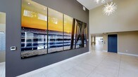 This single-floor space makes a bold first impression through its grand, open entryway or lobby area. The most commanding feature is a large-scale, multi-panel photographic artwork mounted on a charcoal accent wall, depicting a vivid coastal sunset over an ocean pier. The vibrant warm tones of the imagery contrast beautifully against the neutral gray wall, immediately drawing the eye upon entry.

The ceilings soar to an impressive height, amplifying the sense of openness and grandeur throughout the space. Overhead, distinctive starburst chandeliers add a sculptural, contemporary elegance to the interior, serving as decorative focal points that complement the modern aesthetic.

The flooring consists of large-format light-toned tiles laid in a clean grid pattern, contributing to a bright and airy feel. The color palette is refined and cohesive, combining warm whites, soft grays, and subtle beige tones to create a sophisticated ambiance.

A hallway extends beyond the feature wall, leading further into the space, where natural or ambient light filters through, hinting at additional open areas. Overall, the design balances modern minimalism with coastal-inspired artistic elements to create an inviting, stylish environment.