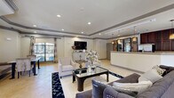 Located at 300 Heron Cove Dr in Enterprise, Alabama, this single-floor space offers a thoughtfully designed open-concept layout that seamlessly connects the living area and kitchen into one cohesive, welcoming environment. The living room is anchored by a striking tray ceiling with gray crown molding detail and recessed lighting, lending a sense of architectural depth and refinement. Large windows dressed with decorative curtains and blinds allow natural light to fill the space, enhancing the warm, neutral color palette found throughout. Light-toned tile flooring runs consistently across the entire floor, contributing to the open and airy feel. A generous seating arrangement, complete with a deep navy sofa, upholstered accent chairs, and a glass-top coffee table, centers the living area with both comfort and style. A dedicated dining nook is visible adjacent to the living room, offering an intimate space for meals. The kitchen is a standout feature, boasting floor-to-ceiling rich dark wood cabinetry, light stone countertops, a white peninsula island, and stainless steel appliances. Pendant lights with warm amber tones hang above the kitchen area, adding character and ambiance. The overall atmosphere is polished and sophisticated, balancing classic design elements with a comfortable, functional layout.