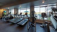 This fitness center is a spacious, single-floor facility designed to support a comprehensive workout experience. The layout is thoughtfully organized, dividing the space into distinct functional zones while maintaining an open, accessible feel throughout. Durable dark rubber flooring covers the entire area, providing both safety and sound absorption during workouts.

The cardio zone is positioned along a wall of large floor-to-ceiling windows, offering unobstructed views of an outdoor pool and surrounding landscape. This area is equipped with multiple treadmills, elliptical trainers, and stationary bikes, allowing users to exercise while enjoying natural light and scenic views. Ceiling fans are mounted overhead to promote airflow and comfort.

The strength training section features a robust selection of selectorized machines, free-weight benches, and a full dumbbell rack. Medicine balls in varying weights are neatly stored on a rack, and mirrored walls in this zone enhance the sense of space while allowing users to monitor their form. A cable machine and additional resistance equipment round out the strength offerings.

The facility is accessible through glass double doors and is well-lit with recessed lighting. Wall-mounted televisions are placed throughout for entertainment. A wellness information board is posted near the entrance, and a sanitizing station is conveniently located at the entry point.