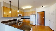 Located at 5137 Gateway St SE in Prior Lake, Minnesota, this single-floor unit offers a well-designed layout that balances functionality and comfort. The kitchen is a standout feature, showcasing rich dark granite countertops that contrast beautifully with warm maple cabinetry. Hardwood-style flooring, pendant lighting, and a full suite of white appliances — including a range, microwave, and refrigerator — make this kitchen both practical and visually appealing. A breakfast bar with an integrated sink connects the kitchen to the adjacent living area, which features soft neutral carpet, wall sconce lighting, and a clean, open feel. The living space opens directly to a private balcony through a sliding glass door flanked by large windows, flooding the room with natural light and offering views of the surrounding community. The bathroom is finished with tile-style flooring, a vanity with maple cabinetry, and accessibility-friendly grab bars, reflecting a thoughtful and practical design. A generously sized walk-in closet with wire shelving provides ample storage. Throughout the unit, the design maintains a consistent warm and neutral palette, creating a cohesive and welcoming atmosphere well-suited to everyday living.