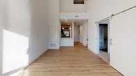 This thoughtfully designed two-floor space offers a compelling blend of modern finishes and open, airy architecture. The main living area immediately impresses with soaring double-height ceilings that amplify the sense of space, complemented by warm-toned hardwood flooring that flows throughout. Large windows invite abundant natural light, casting bright, sweeping patterns across the clean white walls and creating a welcoming atmosphere.

The kitchen is well-equipped with stainless steel appliances, including a gas range with a hood vent and a refrigerator, set against warm wood cabinetry and light-toned granite or stone countertops. Pendant lighting above the kitchen counter adds a contemporary touch.

The bathrooms are finished in a clean, modern style, featuring wood-toned vanities with white countertops, large mirrors, and chrome fixtures. One bathroom includes a soaking tub with shower combination, while another features a sleek, minimalist design with tile flooring.

The bedrooms vary in character — one features soft carpet underfoot with a window overlooking greenery outside, while others offer hardwood-style flooring and clean neutral walls, providing versatile spaces adaptable to various uses. Additional rooms on the second floor provide flexible living or workspace options, maintaining the same bright, understated aesthetic throughout.