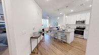 This single-floor residence is designed with a bright, contemporary aesthetic that emphasizes open living and functional design throughout. The heart of the space is an open-concept kitchen and living area, where wood-look flooring flows seamlessly from room to room, enhancing the sense of continuity and space. The kitchen is well-appointed with white shaker-style cabinetry extending to the ceiling, granite countertops, a subway tile backsplash, and a full suite of stainless steel appliances, including a range, microwave, and refrigerator. A central island with seating provides additional prep space and serves as a natural gathering point. Pendant lighting above the island adds a polished finishing touch. The adjoining living area benefits from natural light through large windows fitted with blinds, and a ceiling fan contributes to year-round comfort. The bedroom is a calm, carpeted retreat with generous window placement that invites natural light while maintaining privacy. The bathroom features a glass-enclosed walk-in shower with tile surround, a white vanity with granite countertop, wood-look flooring, and clean, neutral finishes throughout. The overall ambiance is modern, light-filled, and cohesive.