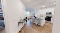 This single-floor residence is designed with a bright, contemporary aesthetic that emphasizes open living and functional design throughout. The heart of the space is an open-concept kitchen and living area, where wood-look flooring flows seamlessly from room to room, enhancing the sense of continuity and space. The kitchen is well-appointed with white shaker-style cabinetry extending to the ceiling, granite countertops, a subway tile backsplash, and a full suite of stainless steel appliances, including a range, microwave, and refrigerator. A central island with seating provides additional prep space and serves as a natural gathering point. Pendant lighting above the island adds a polished finishing touch. The adjoining living area benefits from natural light through large windows fitted with blinds, and a ceiling fan contributes to year-round comfort. The bedroom is a calm, carpeted retreat with generous window placement that invites natural light while maintaining privacy. The bathroom features a glass-enclosed walk-in shower with tile surround, a white vanity with granite countertop, wood-look flooring, and clean, neutral finishes throughout. The overall ambiance is modern, light-filled, and cohesive.