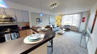 Located in Seattle's vibrant Fremont neighborhood, this thoughtfully designed unit at 620 N 34th St offers a seamless open-concept layout that connects the kitchen, dining, and living areas into one cohesive living space. The kitchen features light wood cabinetry paired with dark countertops, offering a modern contrast that anchors the space. A curved breakfast bar with pendant lighting — including a striking blue accent fixture — serves as both a functional dining surface and a visual centerpiece. The living area is bright and welcoming, benefiting from a large window that frames views of the surrounding Seattle streetscape and fills the room with natural light. Soft, neutral carpeting and contemporary furnishings contribute to a relaxed yet refined ambiance. Track lighting overhead provides additional illumination, complementing the warm glow of floor lamps throughout. The open flow between spaces makes the layout feel expansive and versatile, ideal for both everyday living and entertaining. The design balances modern aesthetics with practical functionality, making this a well-appointed unit in one of Seattle's most walkable and sought-after neighborhoods.