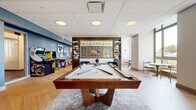 Located at 360 State Street in the heart of New Haven, CT, this resident amenity lounge is a thoughtfully designed recreational space that blends entertainment, comfort, and style into a single open-plan floor. The herringbone-patterned light wood flooring extends throughout, lending a warm, cohesive feel to the entire space.

At the center of the room, a full-size billiard table anchors the entertainment area, surrounded by an ornate area rug. A striking gold geometric room divider with flanking built-in shelving serves as a sophisticated focal point, separating the gaming zone from a cozy dining nook. The dining area features a banquette-style booth with a dark table and plaid accent chairs, bathed in natural light streaming through floor-to-ceiling windows with outdoor views.

The lounge area is defined by bold blue accent walls, a tan leather sofa, jewel-toned blue armchairs, a shuffleboard table, and large-scale artwork. A dedicated arcade corner adds a playful touch with classic stand-up arcade machines. A wall-mounted flat-screen TV and recessed ceiling lighting complete the space. The overall ambiance is modern and energetic, ideal for socializing and recreation.
