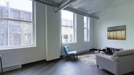 This single-floor space showcases a compelling blend of industrial and modern design elements. The living area immediately draws attention with its notably high ceilings, from which large exposed silver HVAC ductwork runs horizontally, adding an unmistakable industrial character to the space. Crisp white walls provide a clean backdrop that contrasts effectively with the dark-toned hardwood flooring, which runs throughout and grounds the overall aesthetic.

Multiple oversized windows line the walls, allowing generous amounts of natural light to pour in while offering views of the surrounding brick building exterior. The tall windows feature classic double-hung styling, contributing both function and architectural interest. Baseboard heating units are positioned beneath the windows, ensuring year-round comfort.

The living area is open and spacious, providing ample room for furnishings and flexible arrangement. Exposed ceiling beams complement the industrial ductwork overhead, reinforcing the loft-inspired character of the space. The combination of raw architectural details with clean, white-painted surfaces creates a balanced, contemporary atmosphere well-suited for both everyday living and entertaining.