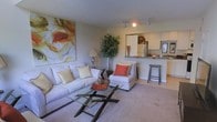 This two-story residence offers a thoughtfully designed interior that balances comfort and functionality. The main living area features plush wall-to-wall carpeting and is anchored by a generously sized sofa and coordinating accent chair, both dressed in neutral tones with colorful throw pillows that add warmth to the space. A glass-top coffee table with a sleek geometric base serves as a stylish focal point, while a large abstract canvas on the wall adds an artistic touch.

The living area flows seamlessly into the kitchen, which is separated by a raised breakfast bar with bar stool seating — ideal for casual dining or entertaining. The kitchen is equipped with stainless steel appliances, white cabinetry, and a window that allows natural light to filter in. Track lighting overhead provides additional illumination across both spaces.

A tall indoor plant near the kitchen transition adds a natural, organic element to the overall aesthetic. The open-concept layout between the living and kitchen areas creates a sense of spaciousness, while the two-floor layout of the residence offers additional living or sleeping space above, providing a well-rounded and versatile home environment.