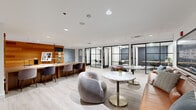 Located at 479-483 N Harlem Ave in Oak Park, IL, this professionally designed coworking suite offers a thoughtfully organized single-floor layout encompassing a generous amount of usable space. The interior is defined by a cohesive modern aesthetic, blending warm cedar wood wall panels with light hardwood-style flooring, crisp white ceilings with recessed lighting, and black-framed glass partitions that create visual openness while maintaining functional separation between zones.

The main open area serves as a flexible hub, featuring a long communal work table with upholstered task chairs, ideal for focused individual work or collaborative sessions. Adjacent to this is a welcoming lounge zone furnished with plush curved sofas, tan leather seating, and marble-topped accent tables on brushed gold bases — providing a relaxed environment for informal meetings or breaks.

Several enclosed private rooms line the perimeter, accessed through sleek black-framed glass doors. These rooms are individually styled, with one featuring geometric patterned wallcovering, offering intimate settings for private calls or small meetings. A dedicated elevator lobby showcases rich wood wall cladding and a framed art piece, lending a polished, gallery-like arrival experience. Curated artwork is displayed throughout, reinforcing a refined, creative atmosphere across the entire suite.