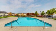 Located at 5701 Ebner Cir in Dublin, Ohio, this outdoor community amenity area centers around a generously sized swimming pool with a classic geometric shape and multiple entry points via stainless steel handrails. The pool is enclosed by a sturdy black iron fence, providing both safety and definition to the space. Surrounding the pool is an expansive concrete pool deck outfitted with numerous blue lounge chairs, offering ample room for sunbathing and relaxation. Adjacent to the pool sits a single-story clubhouse structure featuring a distinctive hip roof with dark shingles, a prominent stone chimney, and large windows that allow natural light to fill the interior. The building's exterior blends neutral tones with white trim detailing, giving it a clean, welcoming appearance. A covered porch area on the clubhouse provides shaded outdoor seating. The grounds beyond the fenced pool area are well-maintained, featuring neatly trimmed green lawns and mature deciduous trees that offer a natural, park-like backdrop. Views of the surrounding residential community are visible from the deck, reinforcing the neighborhood atmosphere. Overall, this amenity space is well-suited for community gatherings and outdoor recreation.