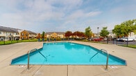 Located at 5701 Ebner Cir in Dublin, Ohio, this outdoor community amenity area centers around a generously sized swimming pool with a classic geometric shape and multiple entry points via stainless steel handrails. The pool is enclosed by a sturdy black iron fence, providing both safety and definition to the space. Surrounding the pool is an expansive concrete pool deck outfitted with numerous blue lounge chairs, offering ample room for sunbathing and relaxation. Adjacent to the pool sits a single-story clubhouse structure featuring a distinctive hip roof with dark shingles, a prominent stone chimney, and large windows that allow natural light to fill the interior. The building's exterior blends neutral tones with white trim detailing, giving it a clean, welcoming appearance. A covered porch area on the clubhouse provides shaded outdoor seating. The grounds beyond the fenced pool area are well-maintained, featuring neatly trimmed green lawns and mature deciduous trees that offer a natural, park-like backdrop. Views of the surrounding residential community are visible from the deck, reinforcing the neighborhood atmosphere. Overall, this amenity space is well-suited for community gatherings and outdoor recreation.